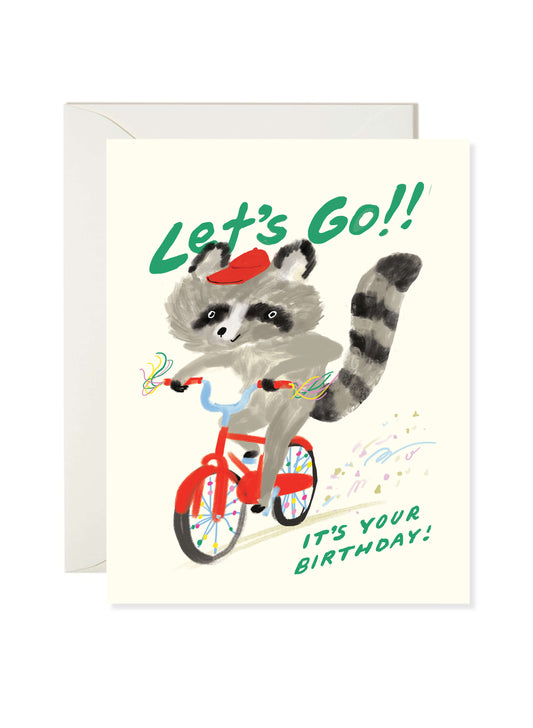 Illustrated birthday greeting card featuring a raccoon riding a red bike with streamers and confetti under “Let’s Go!! It’s Your Birthday!” text by Karen Schipper.