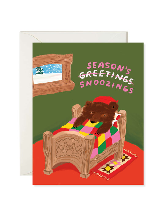 Illustrated Christmas greeting card featuring sleepy bears under a colorful quilt inside a cozy cabin with “Season’s Greetings Snoozings” text by Karen Schipper.