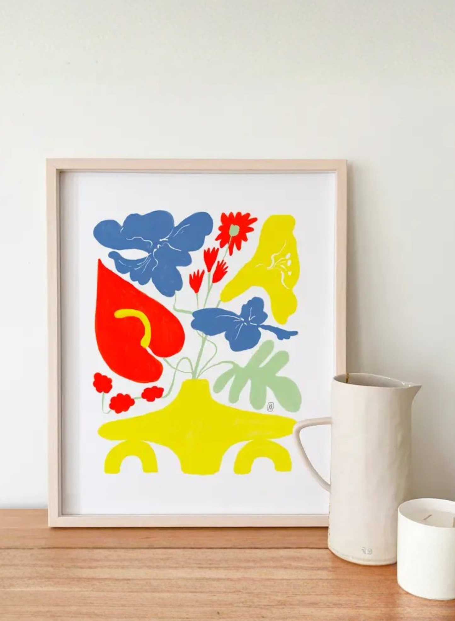 Framed Forsythia Vase Art Print by Karen Schipper styled on a wooden shelf beside a white mug, showing colorful floral wall art in a home decor setting.