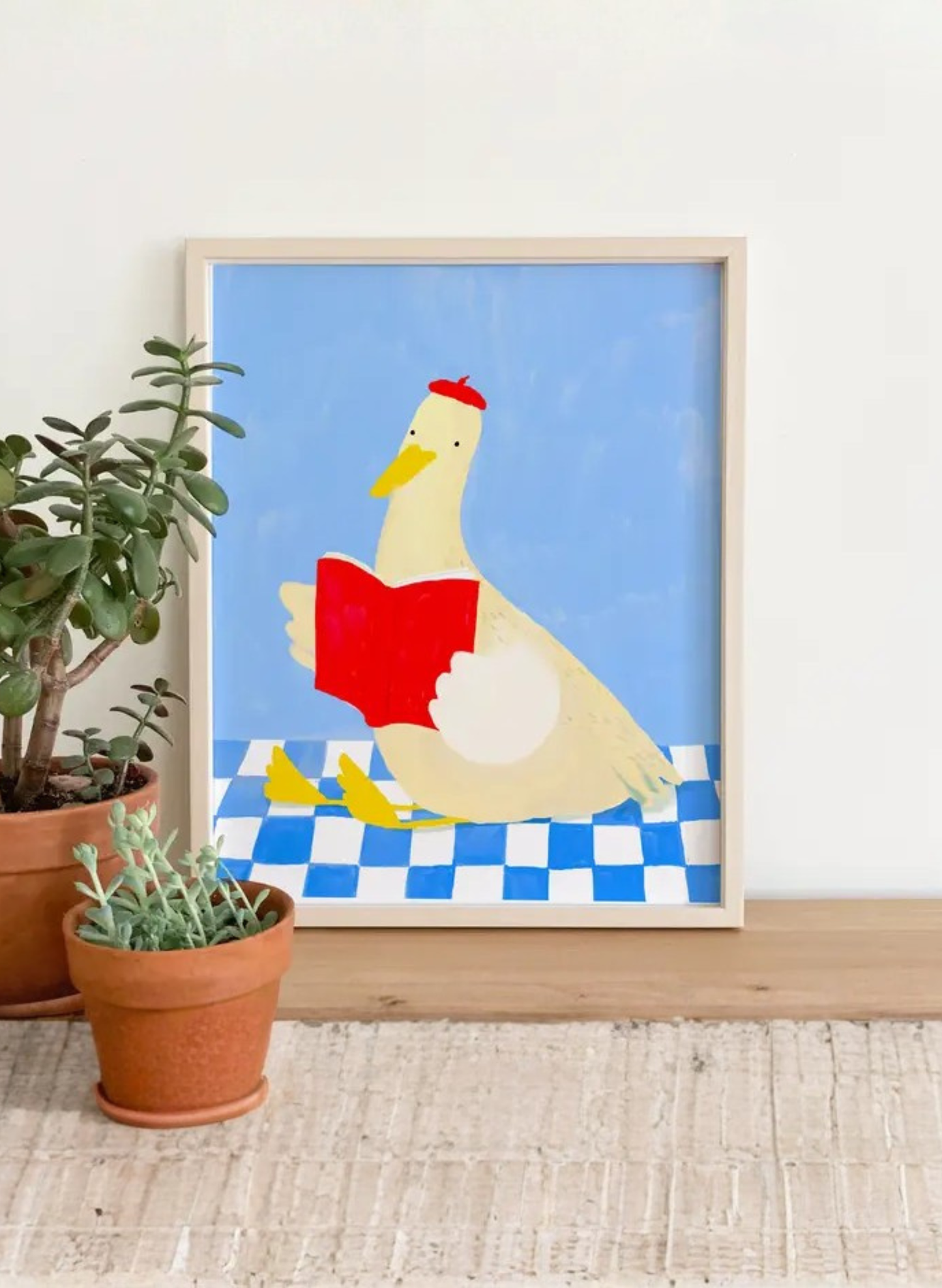 Framed Storytime Duck Art Print by Karen Schipper styled beside potted plants, featuring a duck in a red beret reading a red book on a blue background.