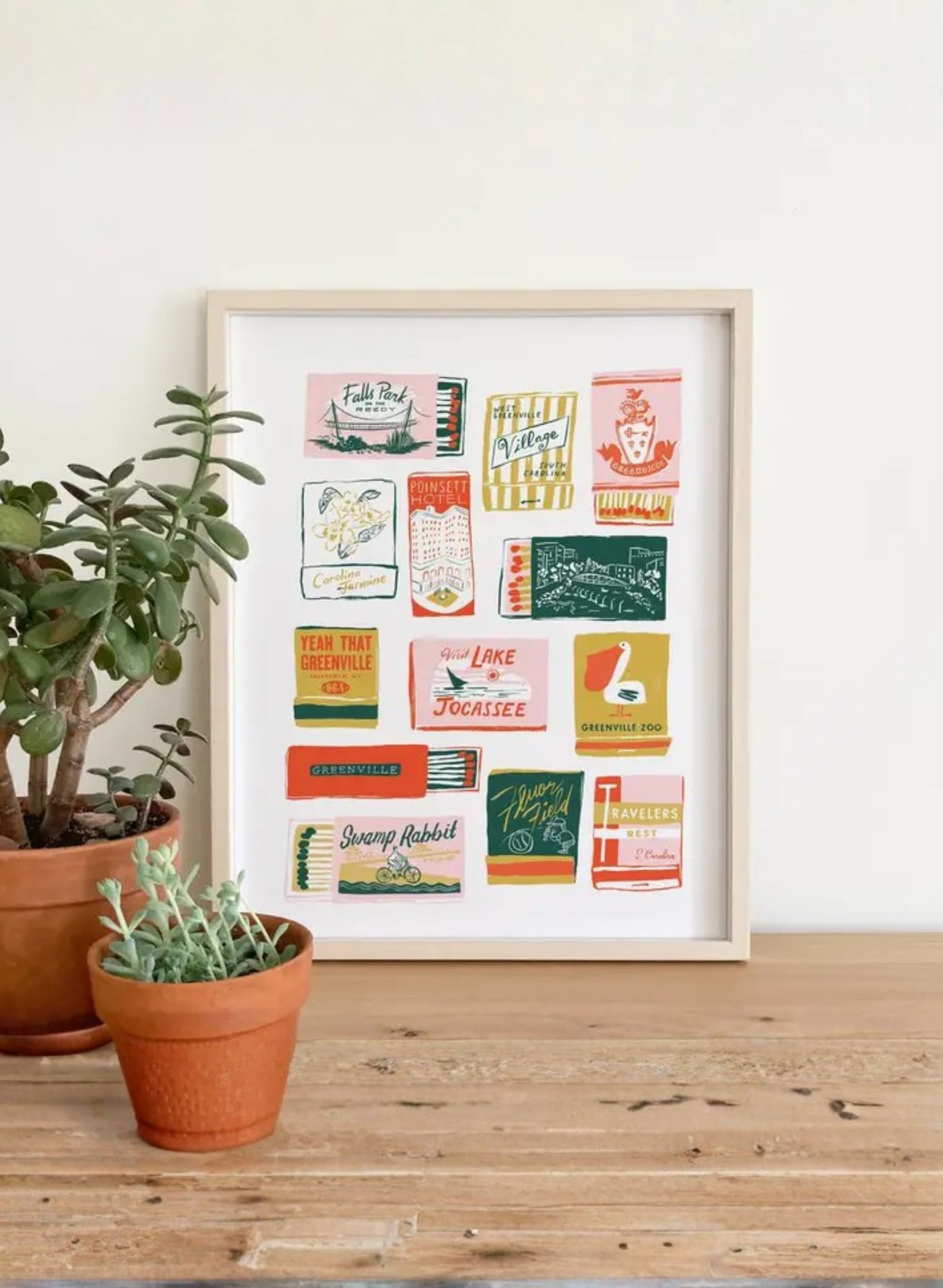 Framed Greenville Matchbooks Art Print by Karen Schipper styled beside potted plants, showing colorful South Carolina city wall art.