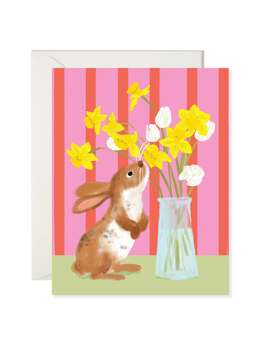 Bunny Spring Bouquet Card by Karen Schipper featuring a brown rabbit beside a vase of yellow daffodils and white flowers on a pink striped background.
