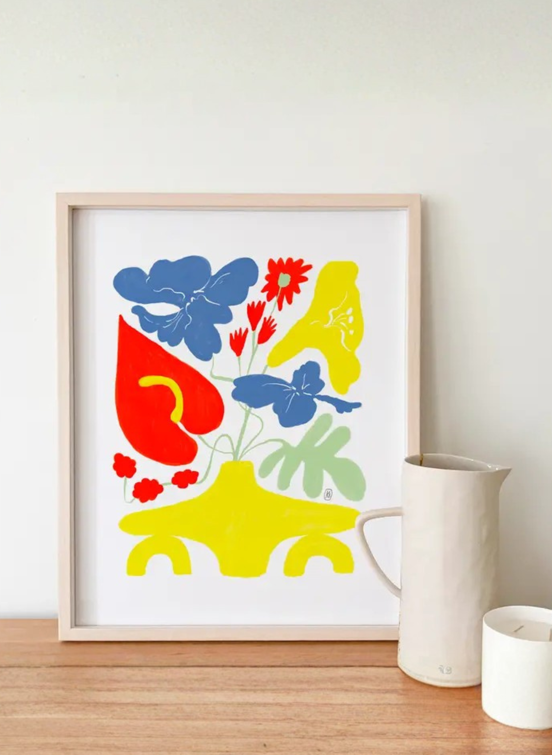 Framed Forsythia Vase Art Print by Karen Schipper styled on a wooden shelf beside a white mug, showing colorful floral wall art in a home decor setting.