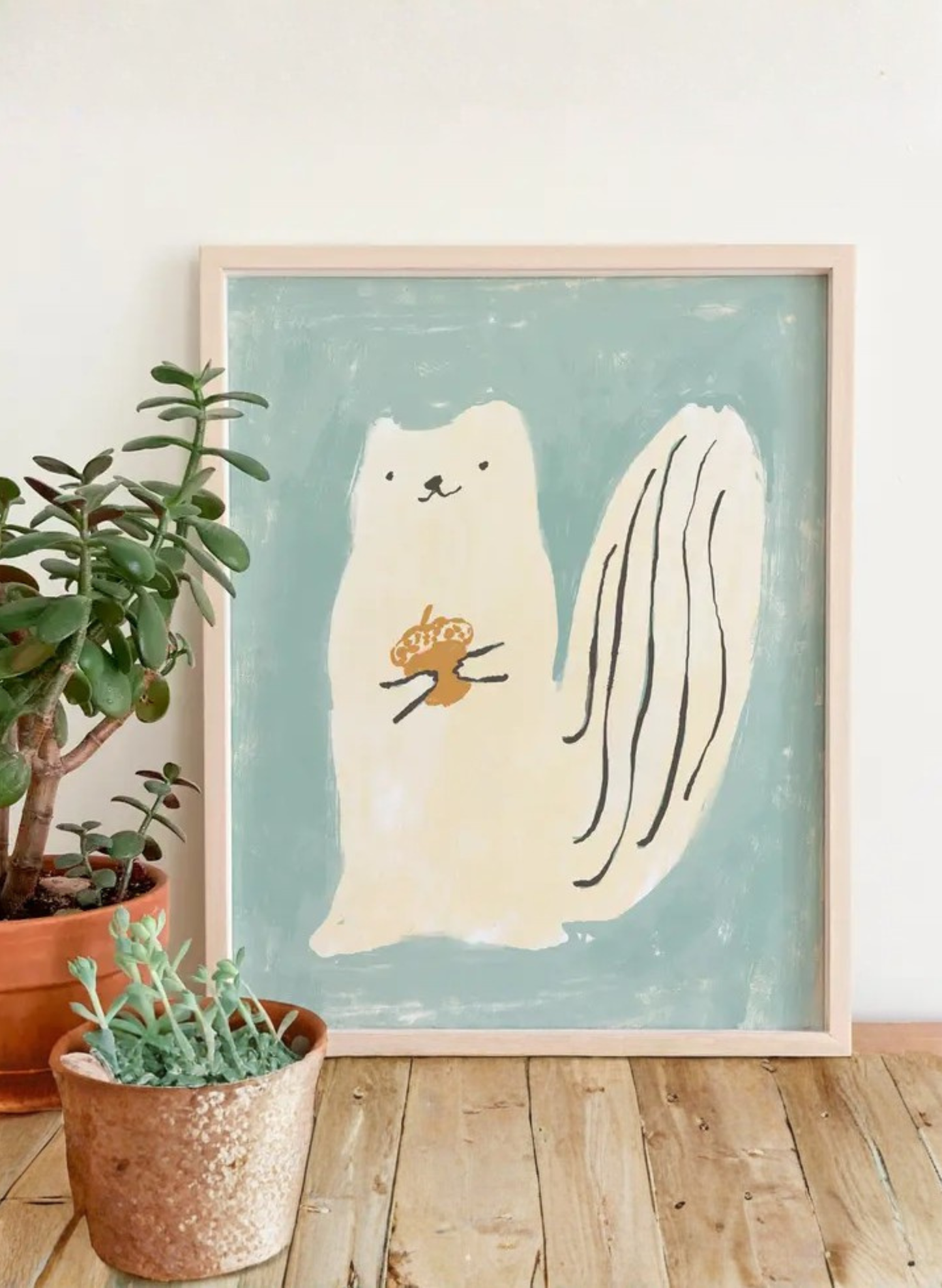 Framed Squirrel Luck Art Print by Karen Schipper styled beside potted plants, showing a cream squirrel holding an acorn on a soft blue-green background.