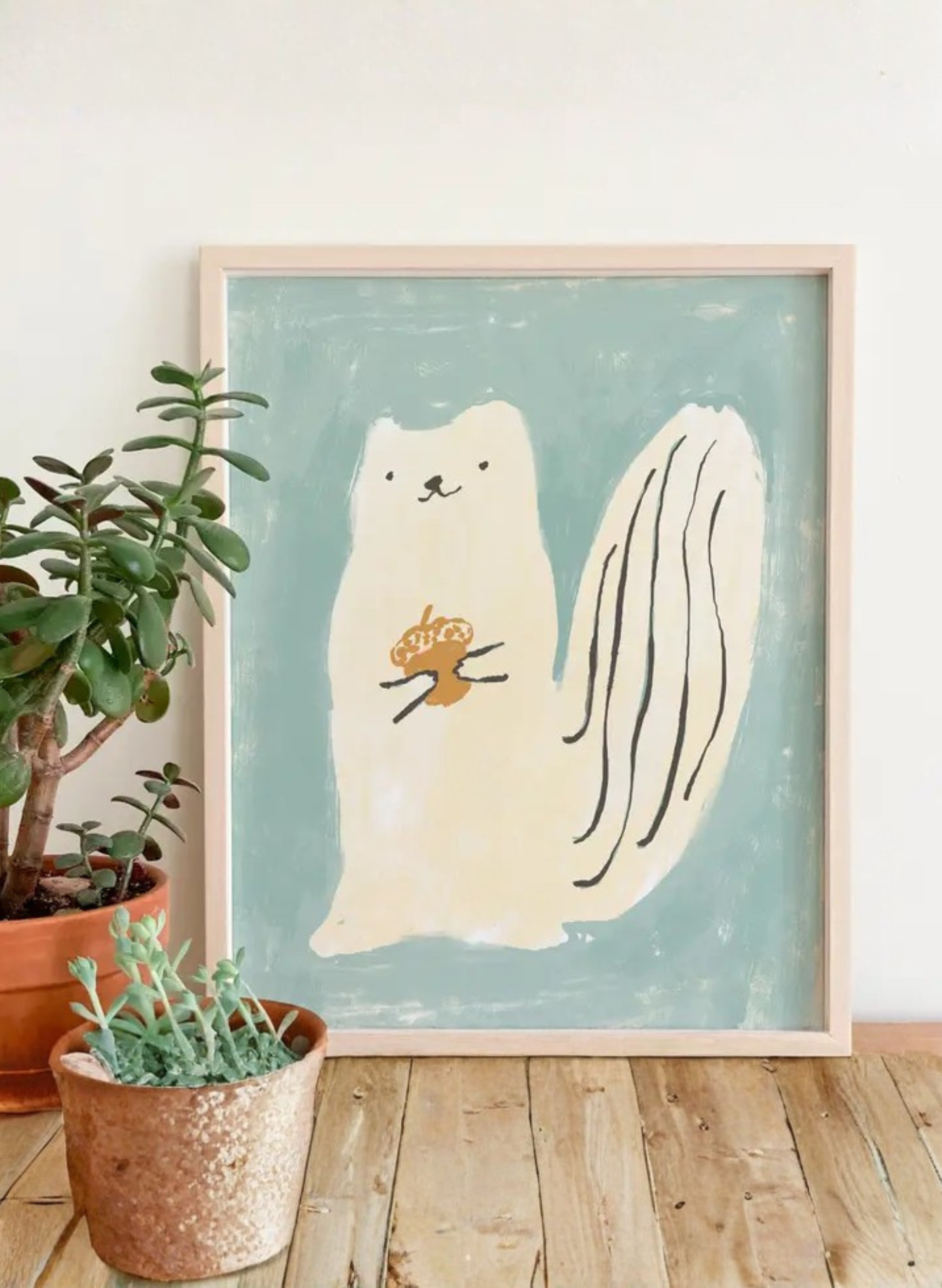 Framed Squirrel Luck Art Print by Karen Schipper styled beside potted plants, showing a cream squirrel holding an acorn on a soft blue-green background.