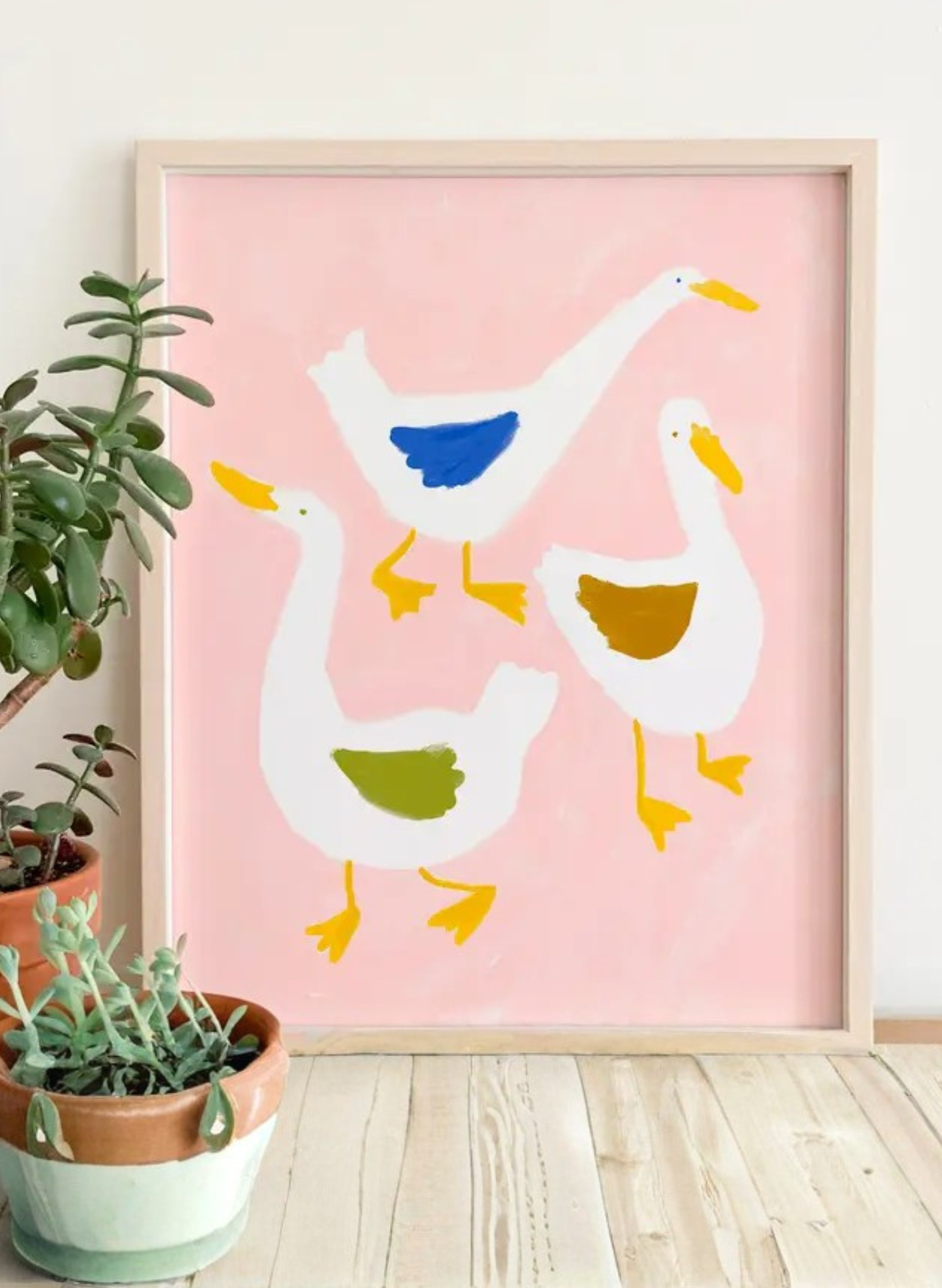 Framed Aylesbury Duck Art Print by Karen Schipper showing three white ducks with colorful wings on a blush pink background styled beside potted plants.