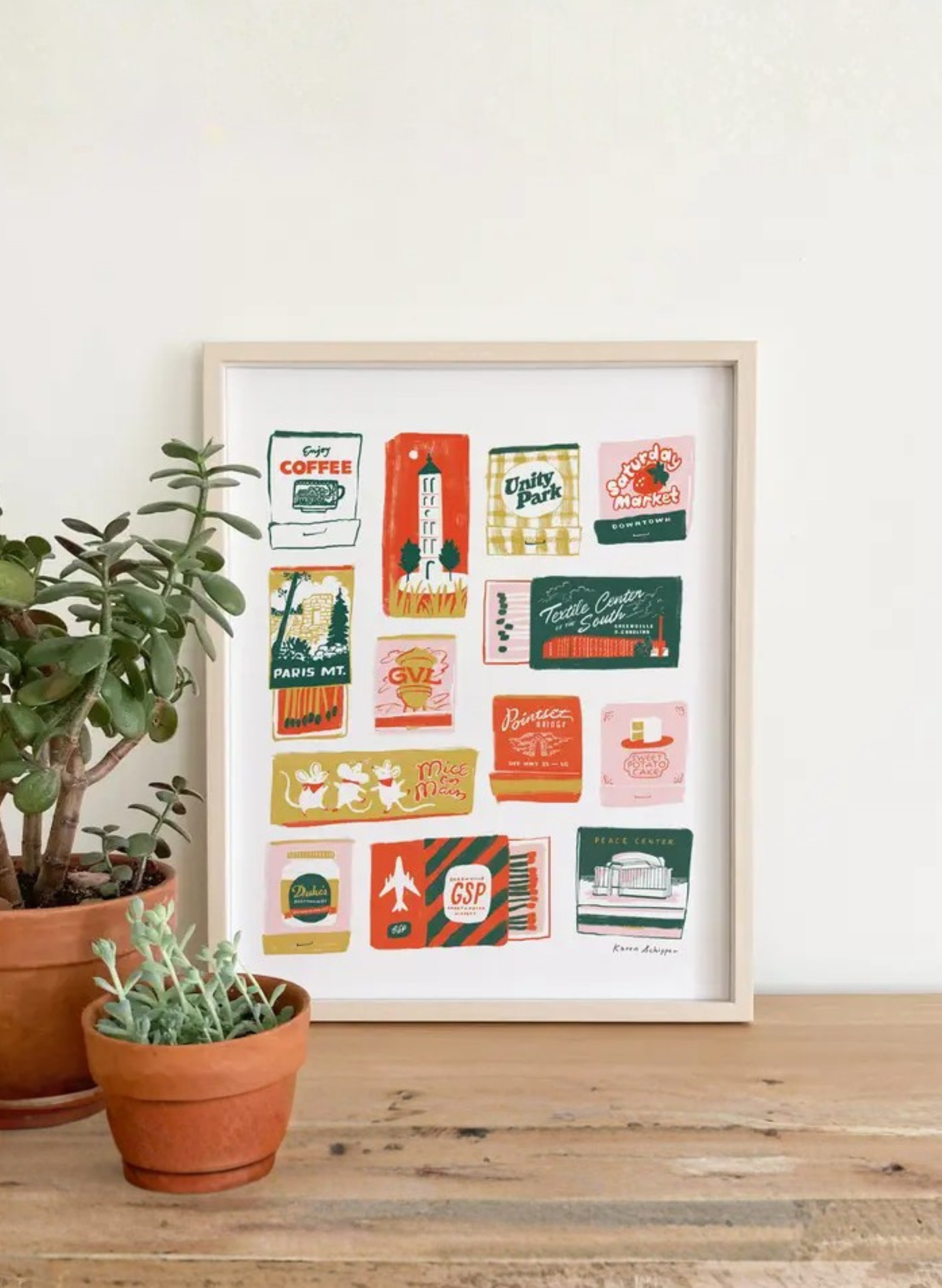Framed Greenville Matchboxes 2.0 Art Print by Karen Schipper styled beside potted plants, featuring colorful South Carolina matchbook-style city artwork.