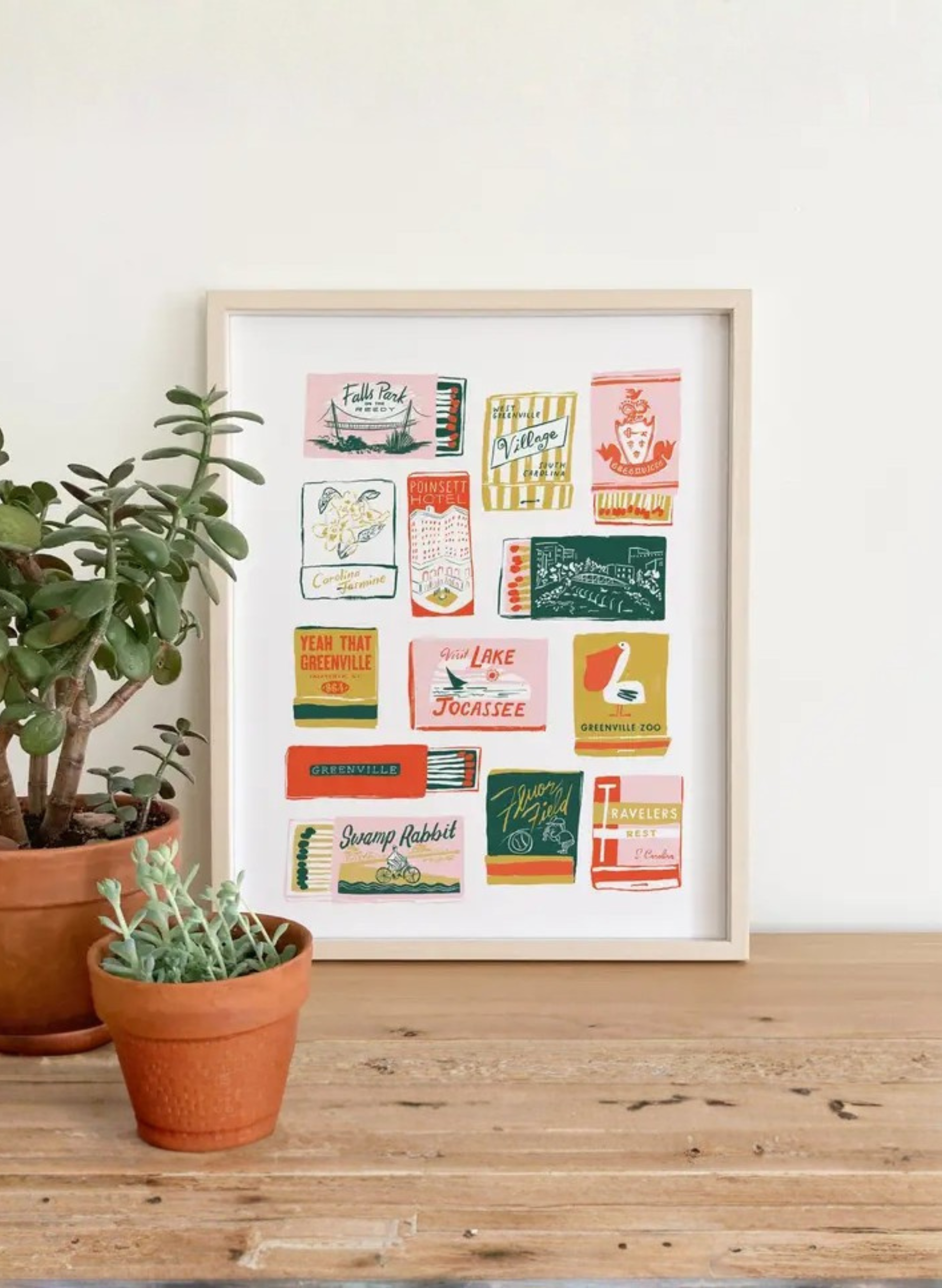 Framed Greenville Matchbooks Art Print by Karen Schipper styled beside potted plants, showing colorful South Carolina city wall art.