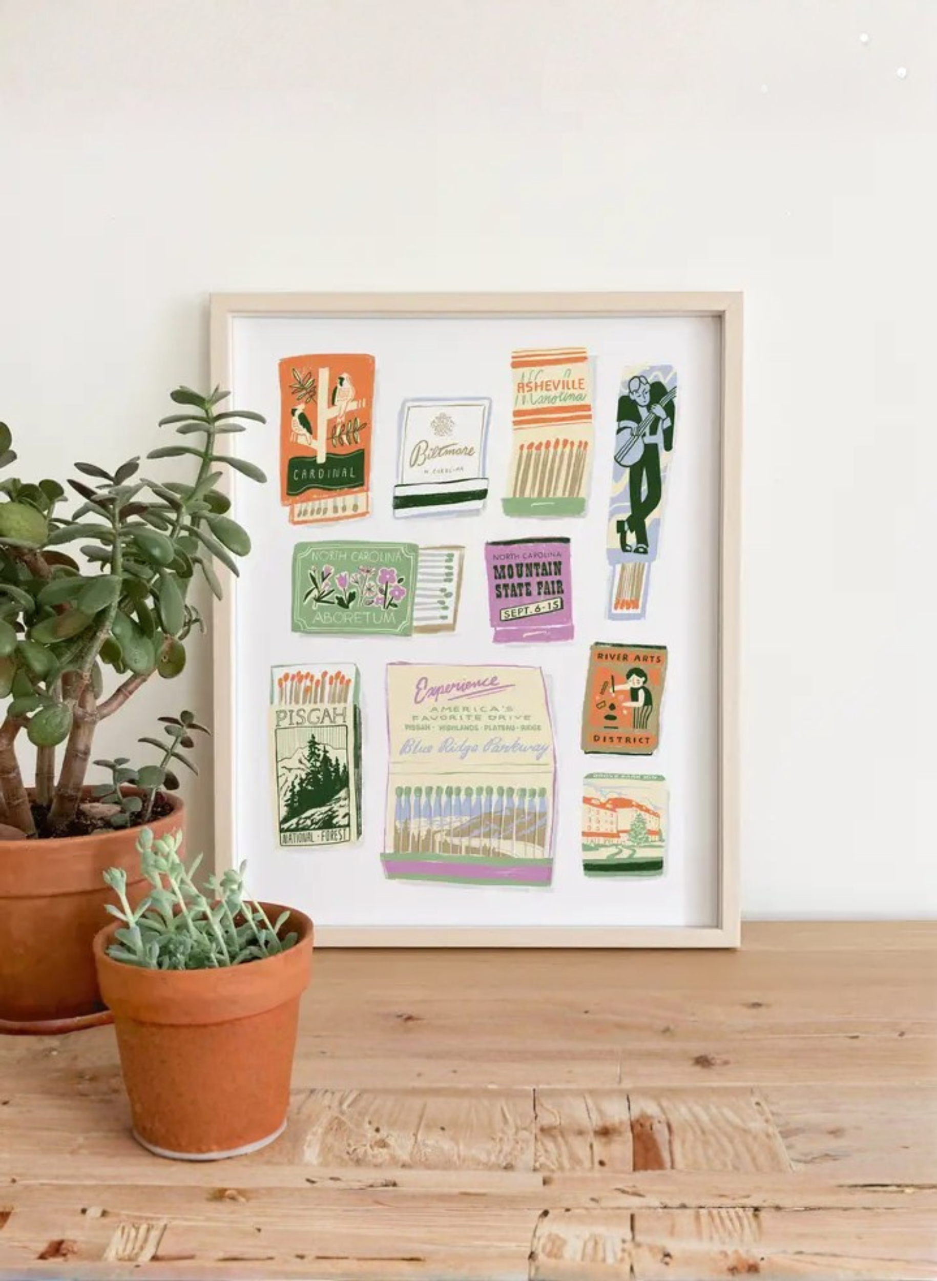 Framed Asheville Matchbook Art Print by Karen Schipper styled beside potted plants, featuring colorful North Carolina matchbook-style landmark artwork.