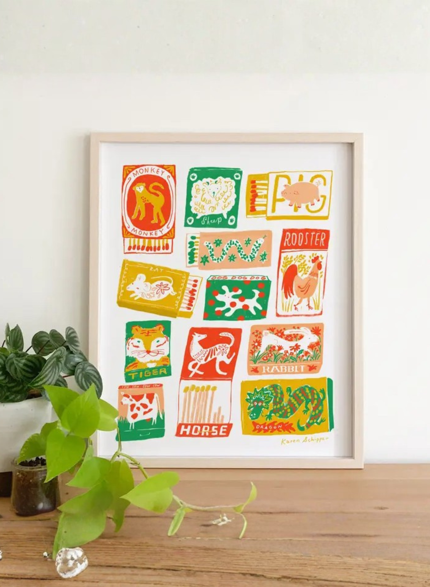 Framed Chinese Zodiac Matchbook Art Print by Karen Schipper styled beside plants, featuring vintage-style matchboxes for the zodiac animals.