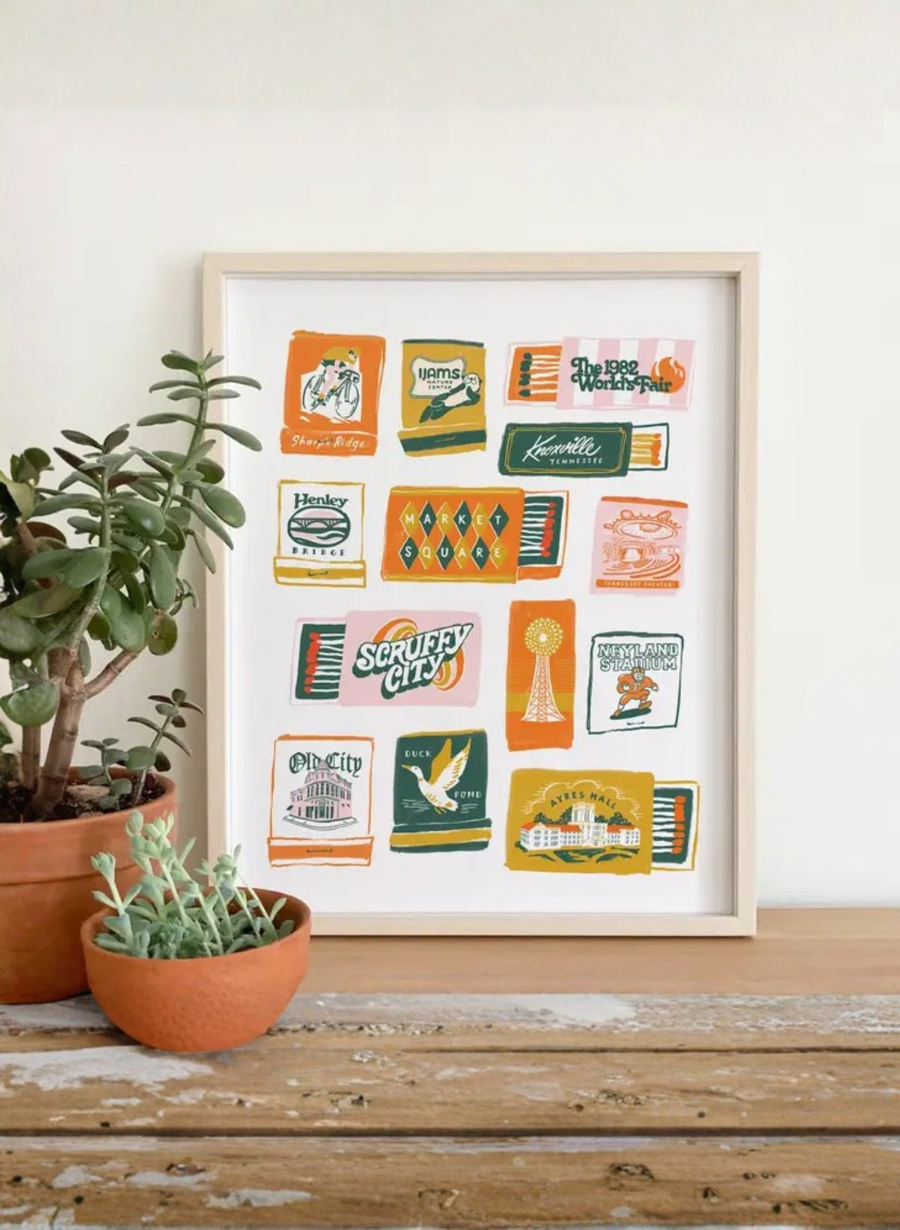 Framed Knoxville Matchbook Art Print by Karen Schipper styled beside potted plants, featuring colorful Tennessee matchbook-style landmark artwork.