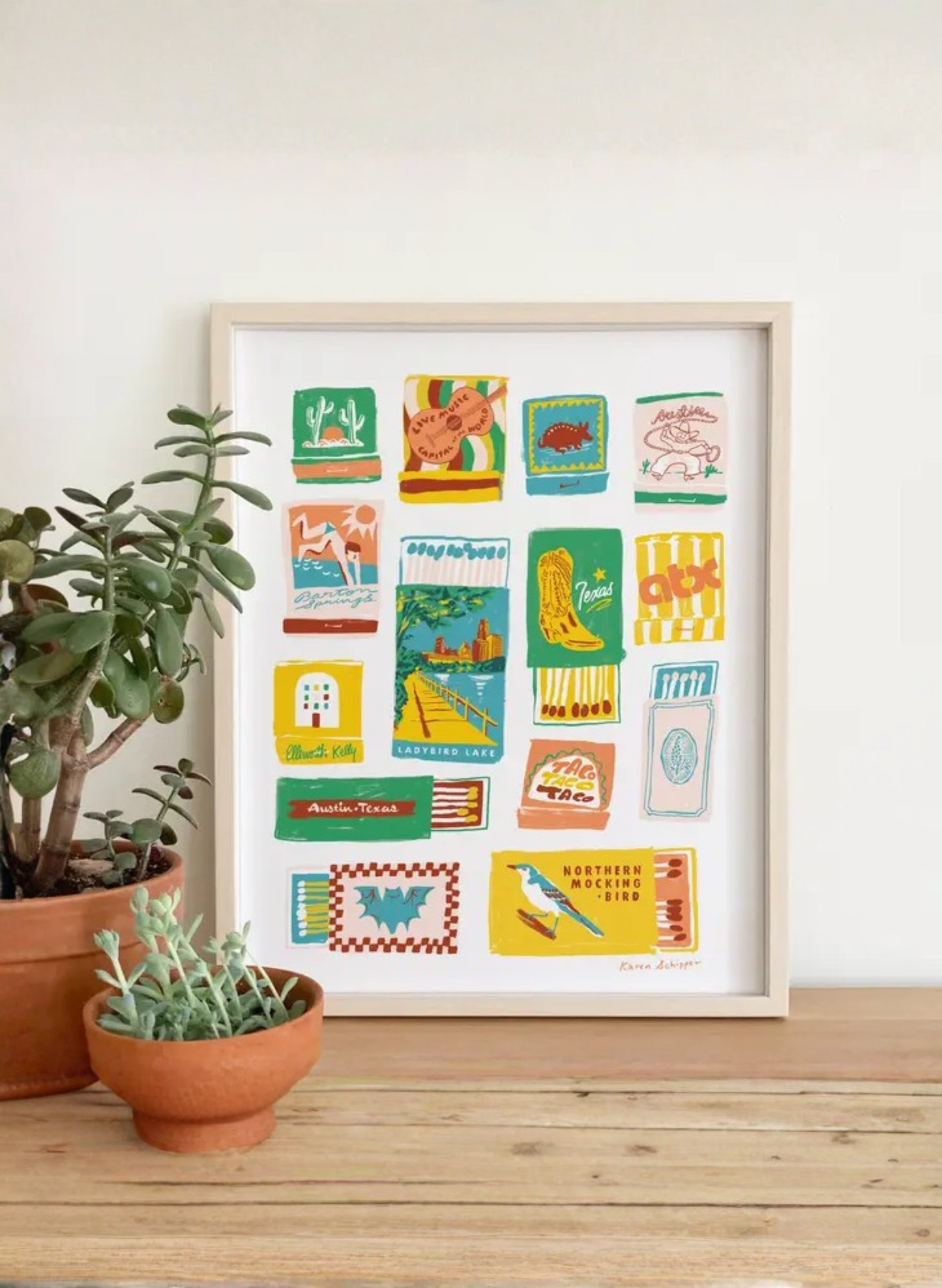 Framed Austin Matchbooks Art Print by Karen Schipper styled on a wooden table with potted plants, featuring vintage-inspired Austin matchbook illustrations and Texas landmarks.