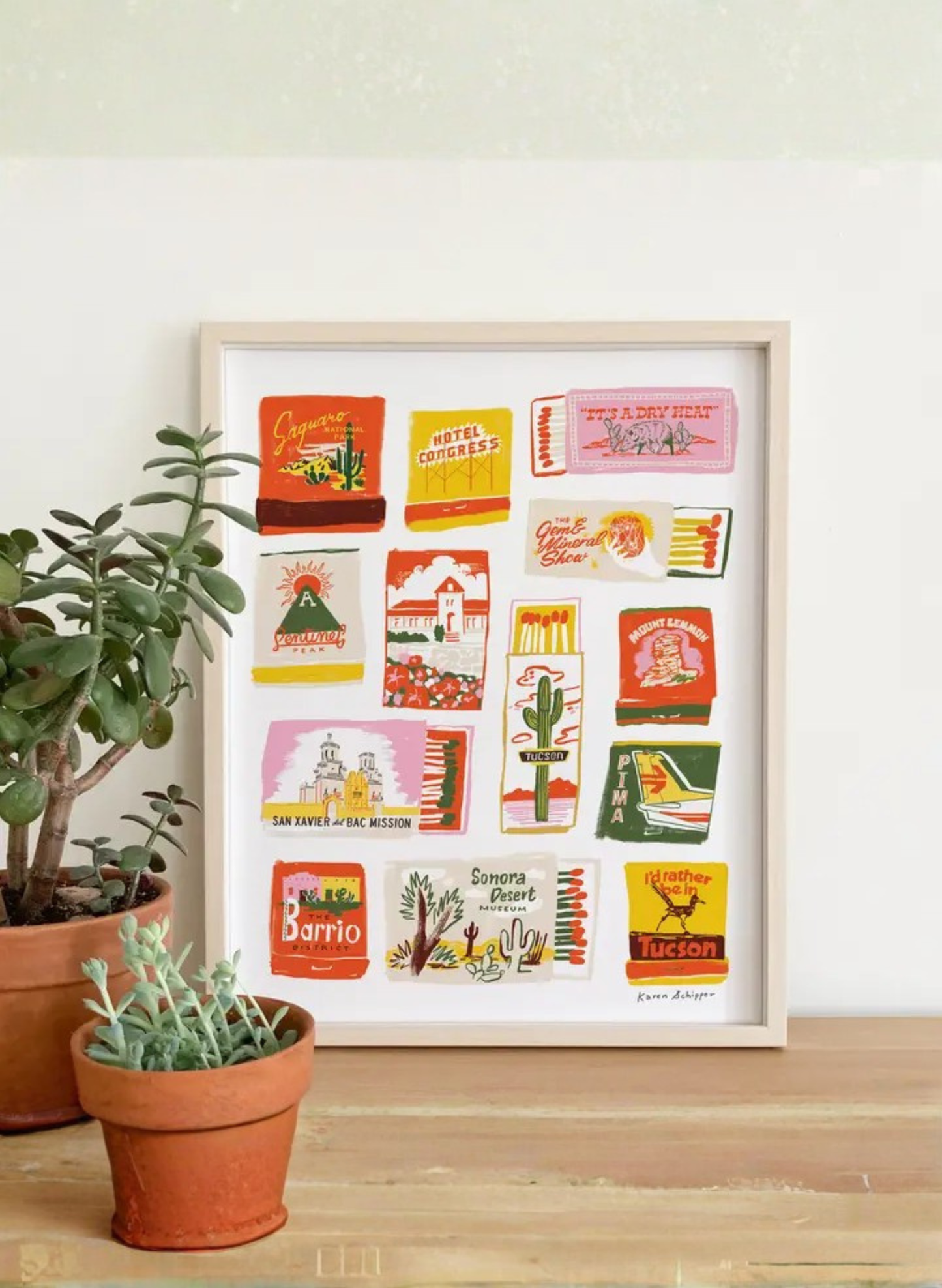 Framed Tucson Matchbook Art Print by Karen Schipper styled beside potted plants, showing colorful Arizona desert wall art and vintage matchbook details.