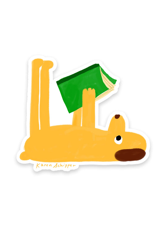 Reading Dog Sticker by Karen Schipper featuring a yellow dog lying on its back holding a green book, illustrated in a simple, playful style.